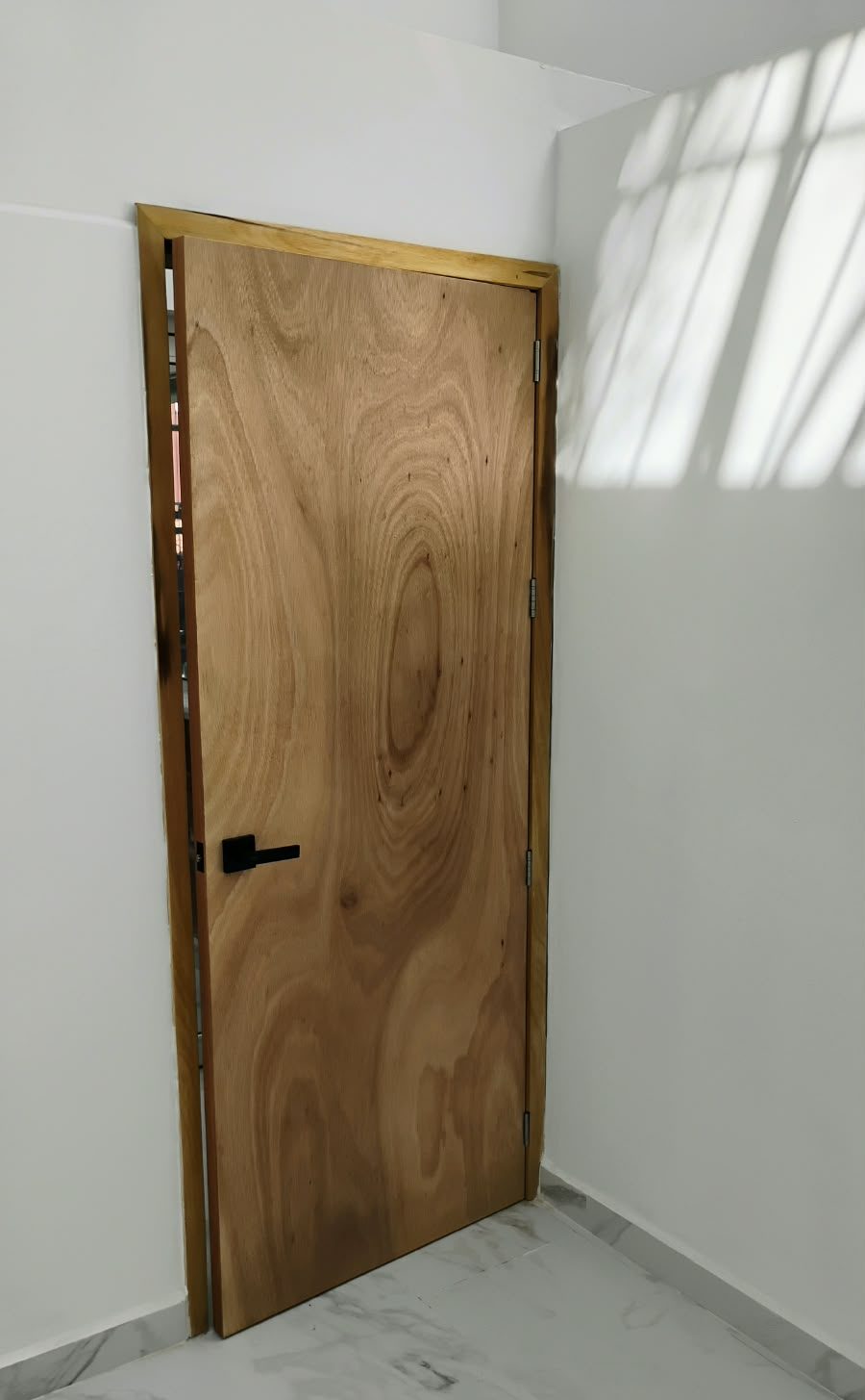 Puerta de madera natural con marco a juego y herraje negro en un cuarto con piso de mármol